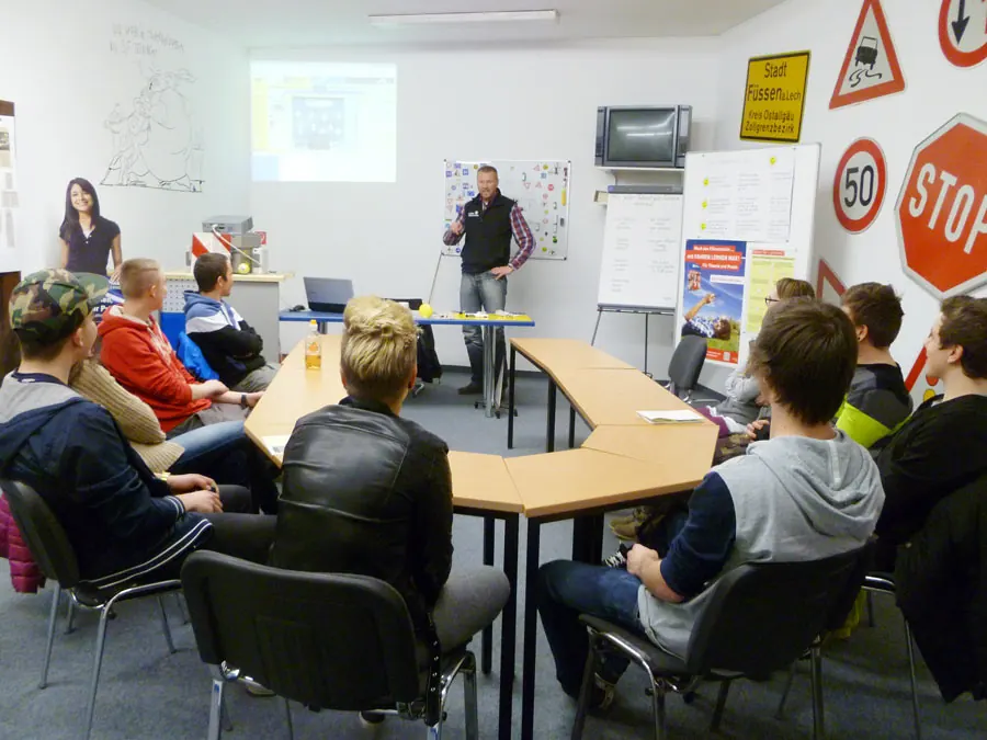 Theorieunterricht in Andi's Fahrschule: Fahrschülerinnen und Fahrschüler an U-förmig gestellten Tischen, vorne der Fahrlehrer an der Leinwand