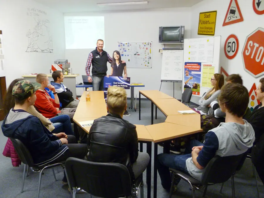 Fahrlehrer erklärt vor der Gruppe den Unterrichtsstoff, die Fahrschüler sitzen an U-förmig gestellten Tischen